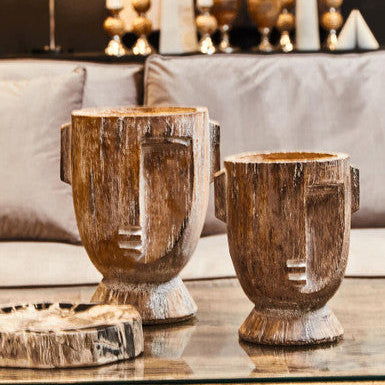 Two rustic wooden vases on a glass table with a blurred background of decorative items.