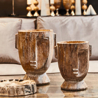 Two rustic wooden vases on a glass table with a blurred background of decorative items.
