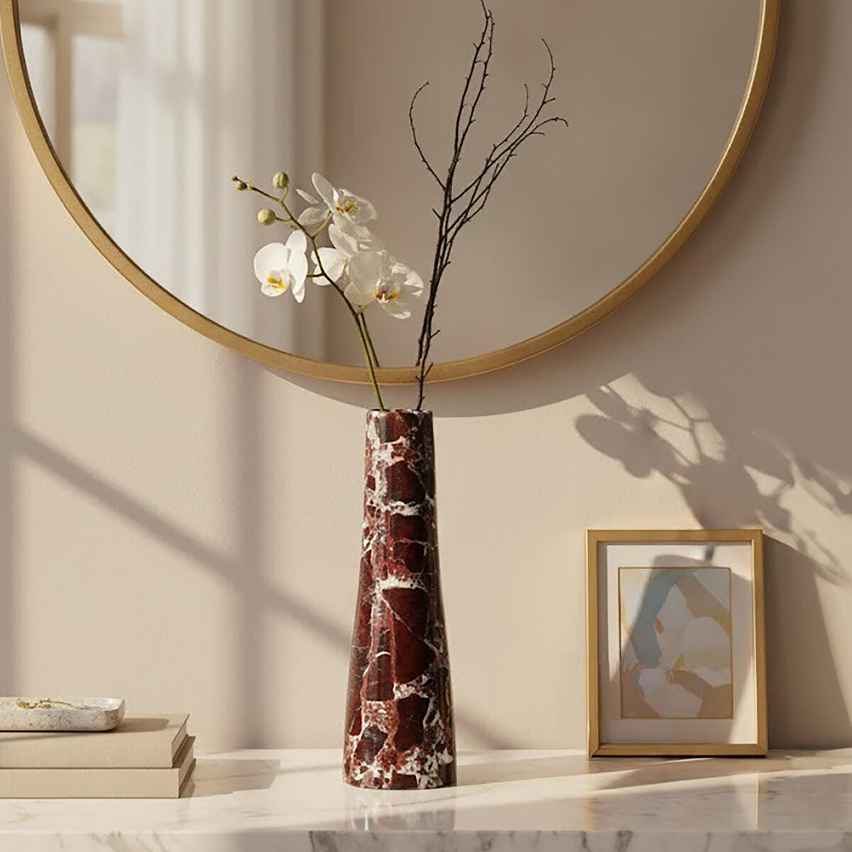 Decorative vase with a white orchid and branch on a marble surface, with a round mirror and abstract art in the background.
