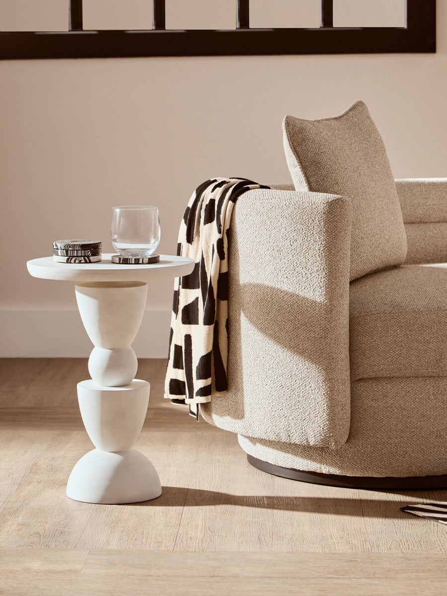 Beige armchair with a patterned blanket next to a white side table in a room.