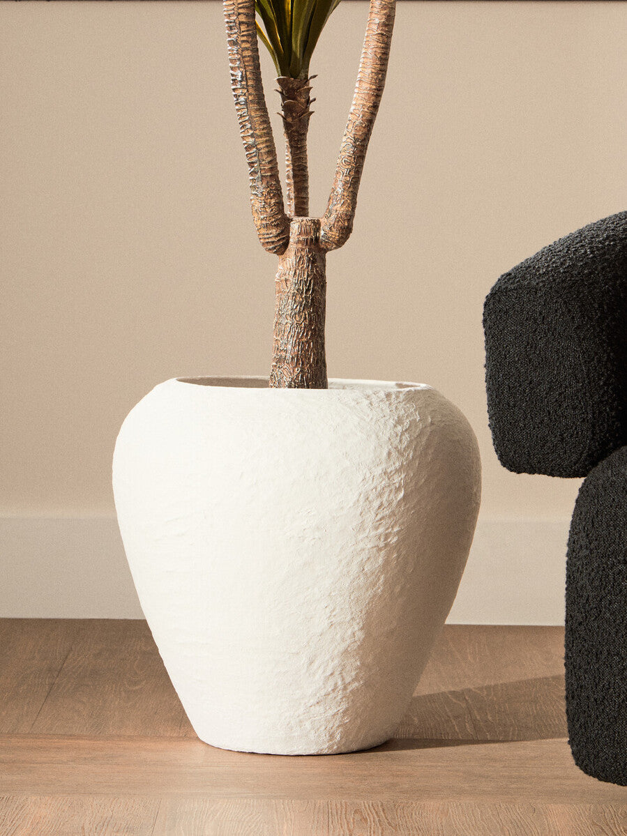 White textured planter with a plant on a wooden floor against a beige wall.