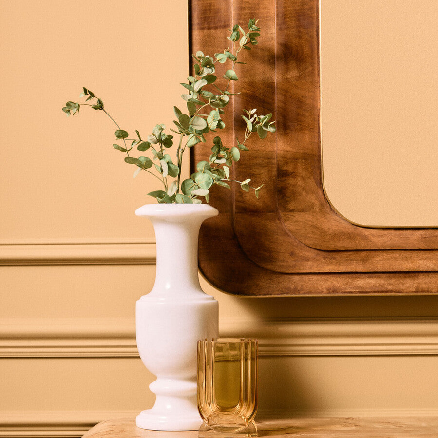 White vase with greenery on a wooden surface against a beige wall.