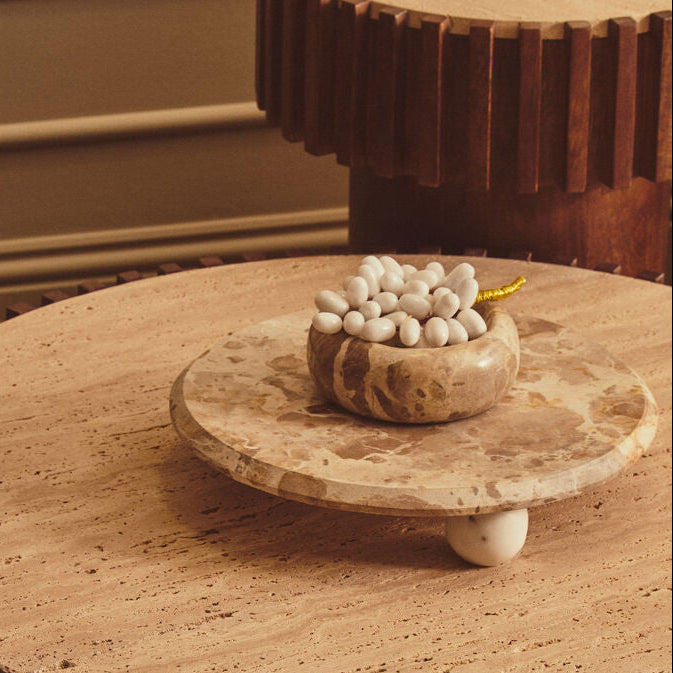 A round coffee table with a fluted dark wood base and a light beige travertine stone top, styled with a small marble bowl of white decorative stones in a warm, elegant interior setting.