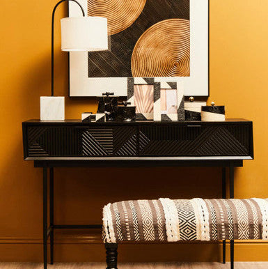 Modern interior design with a black console table, abstract art, and patterned stool against a yellow wall.