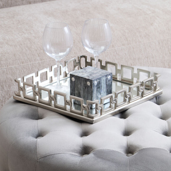 Gray tufted ottoman with a decorative tray and glasses on top, against a neutral background.