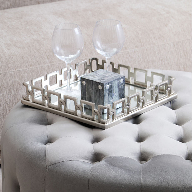 Gray tufted ottoman with a decorative tray and glasses on top, against a neutral background.