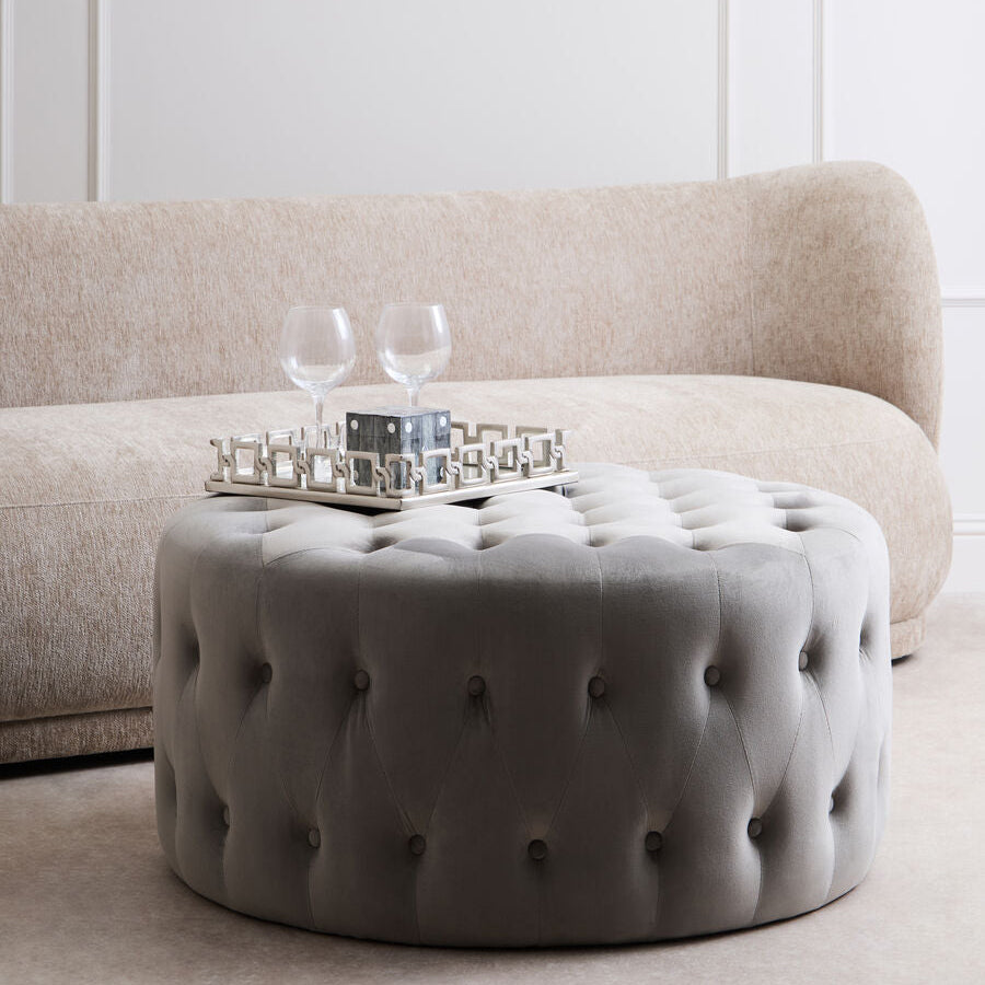 Round tufted ottoman in front of a beige sofa with a white wall background