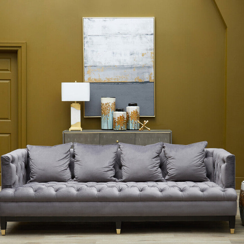 Gray tufted sofa in a room with a painting, lamp, and decorative items on a wooden floor.