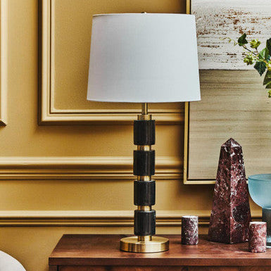 Table lamp with a white shade on a wooden side table against a gold wall.