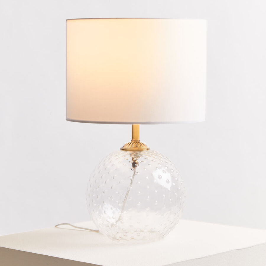 Table lamp with a clear glass base and beige shade on a white surface.