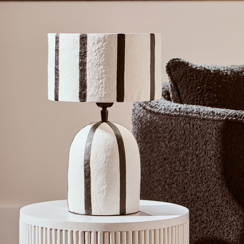 Decorative lamp with striped design on a white side table next to a textured brown chair.