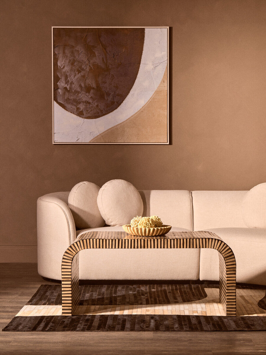 Modern living room with a beige sofa, wicker coffee table, and abstract art on a brown wall.