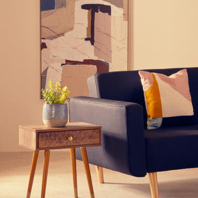 Navy blue sofa with colorful cushions next to a wooden side table with a plant, in a room with abstract art on the wall.