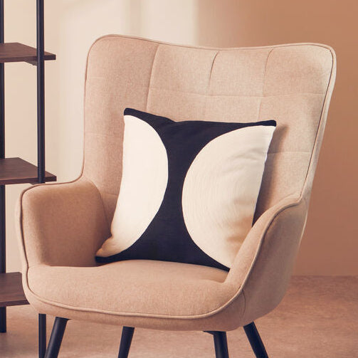 Beige armchair with a decorative pillow next to a wooden shelf against a beige wall.