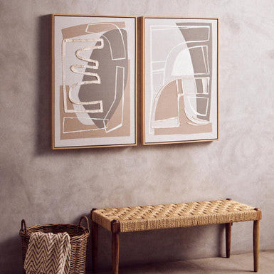 Two abstract framed artworks on a wall with a woven bench and basket below.