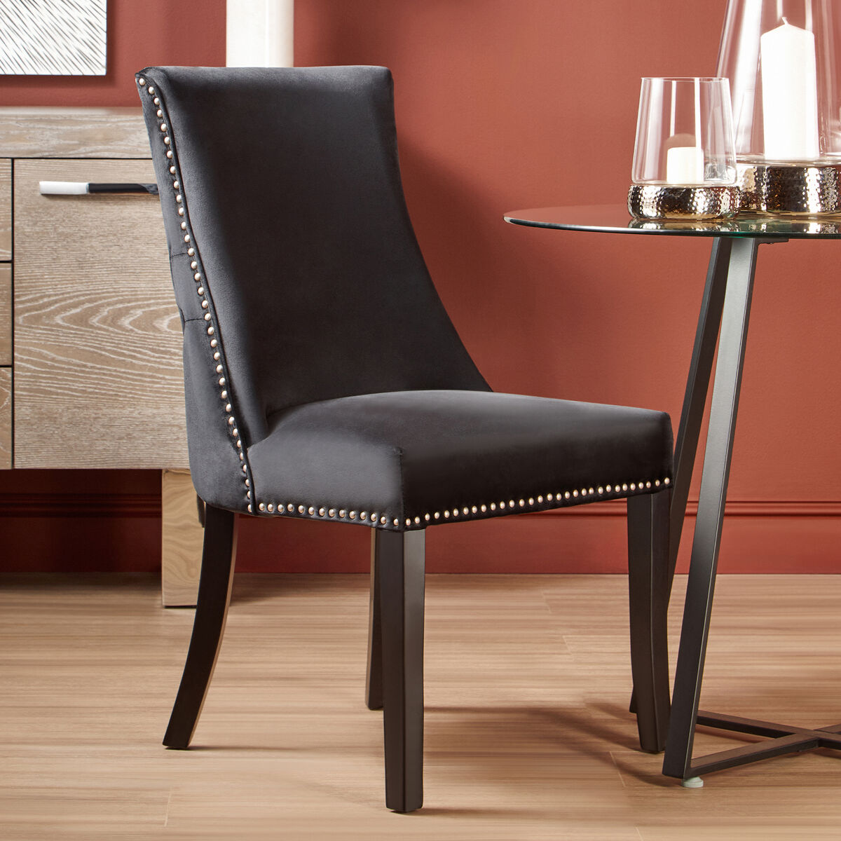 Gray upholstered chair with nailhead trim in a room with a red wall and glass table.