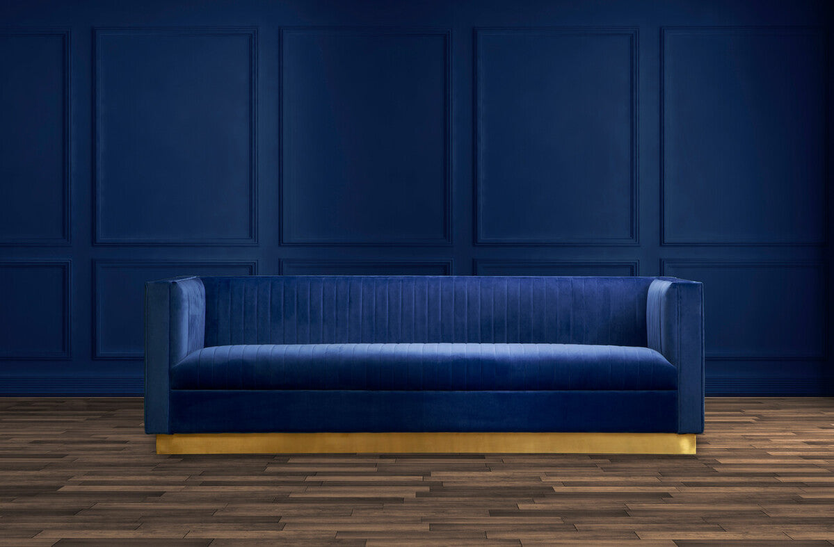 Blue velvet sofa with gold legs against a dark blue paneled wall.