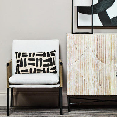 Chair with a white cushion featuring black geometric patterns, next to a wooden cabinet and abstract art.