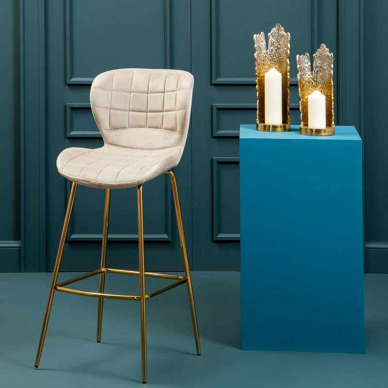 Beige bar stool with gold legs next to a blue pedestal with decorative candles against a teal paneled wall.