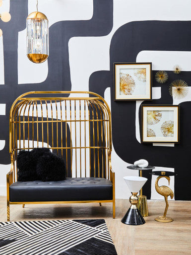 Gold cage chair with black cushion against a black and white patterned wall with framed art.