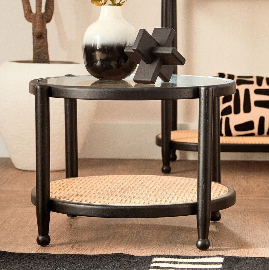Round black side table with a glass top and wicker shelf, featuring decorative items.
