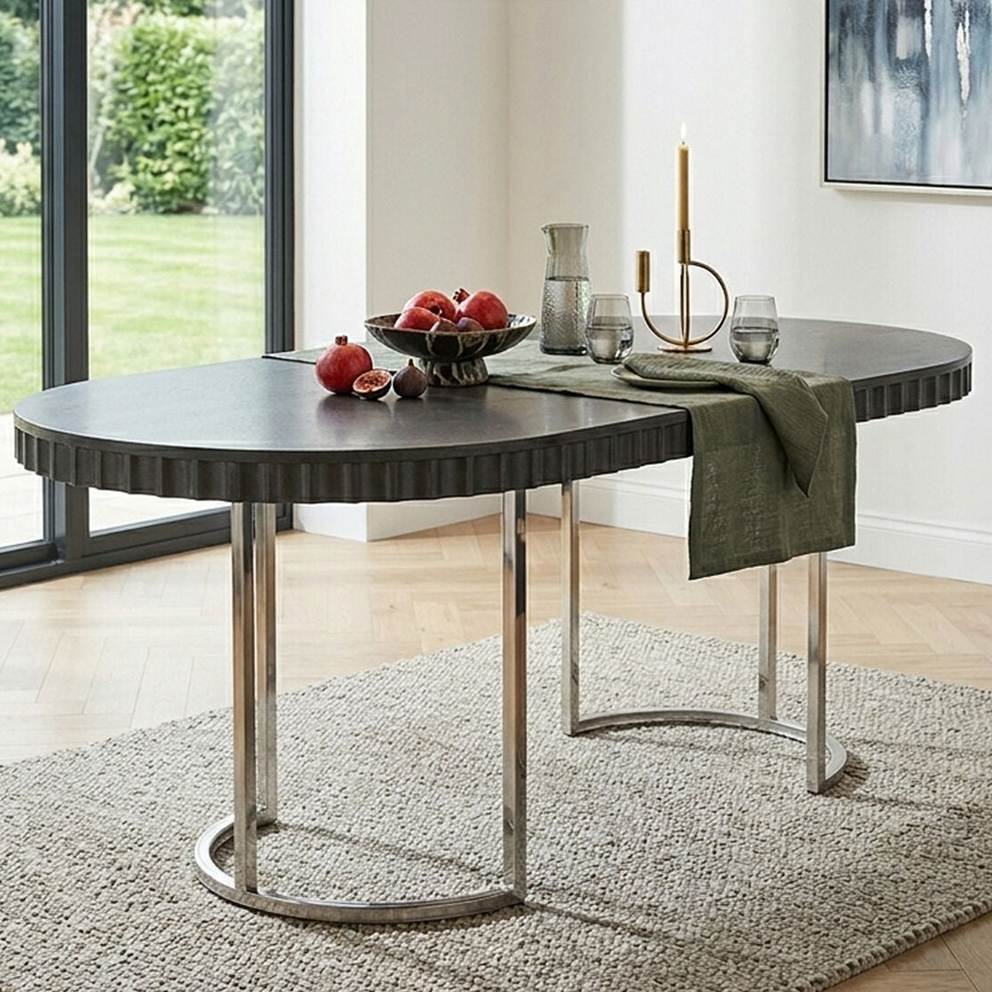 An oval dining table with a dark gray ribbed top and polished silver metal legs, styled in a bright modern dining room with a green throw blanket, fruit bowl, and glassware.