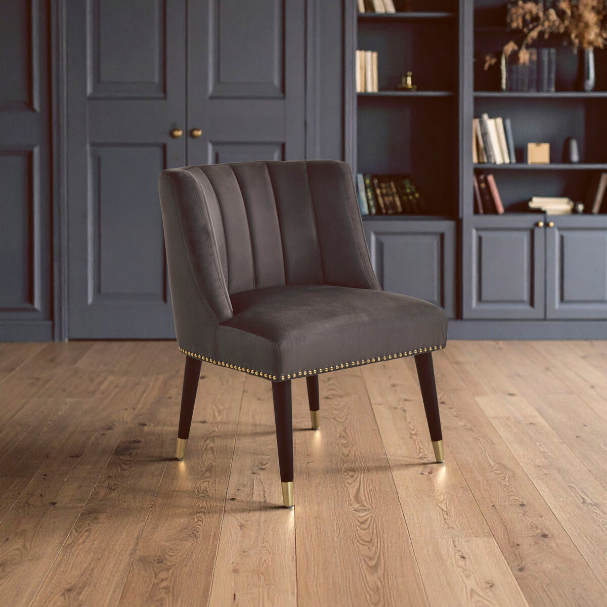 Gray upholstered chair with gold legs on a wooden floor in a room with gray paneling and bookshelves.
