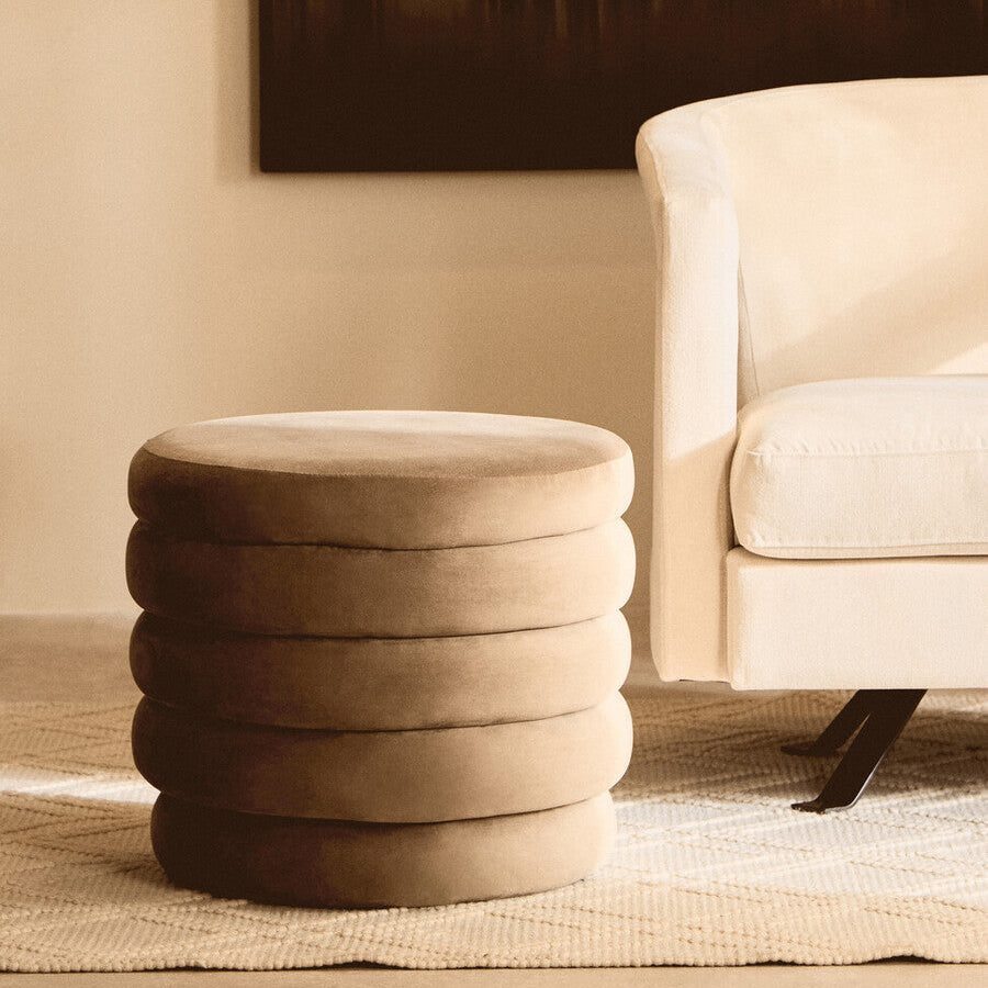 Round cushioned stool in a living room setting with a beige sofa and abstract painting.