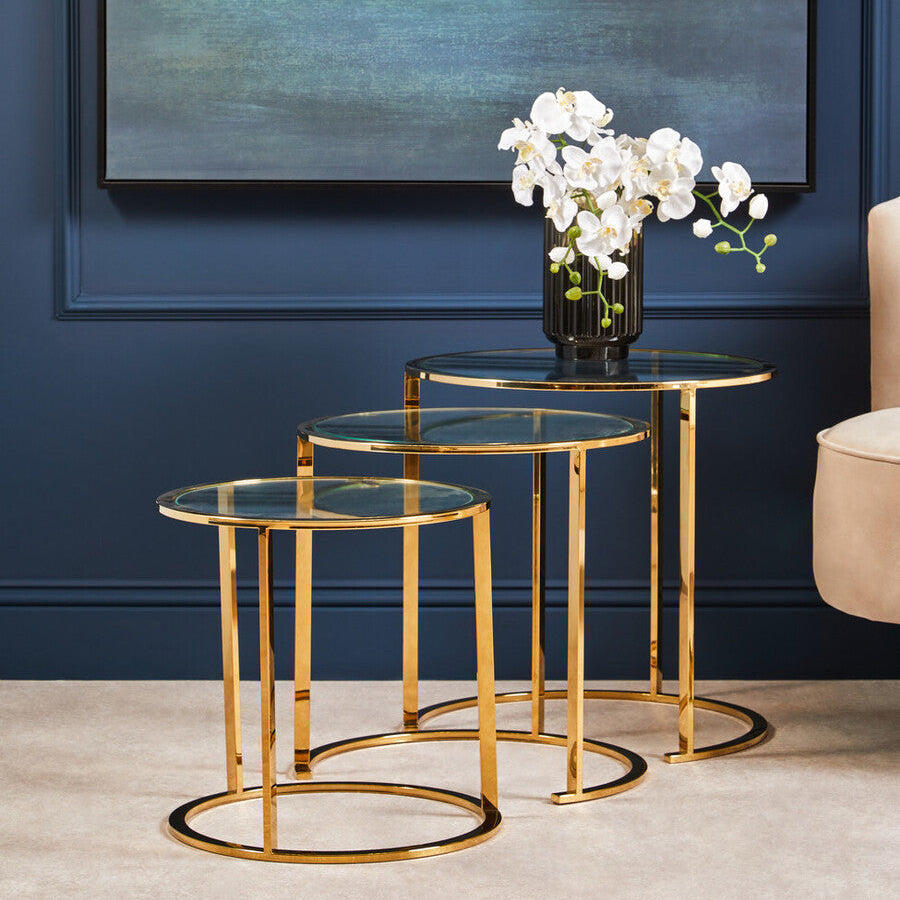 Set of gold nesting tables with glass tops against a blue wall with abstract painting