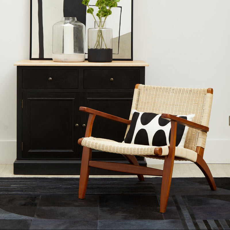 Wicker chair with wooden armrests and a black and white cushion in front of a black cabinet.