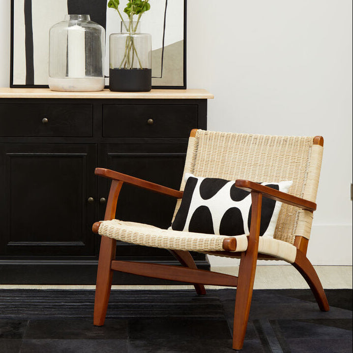 Wicker chair with wooden armrests and a black and white cushion in front of a black cabinet.