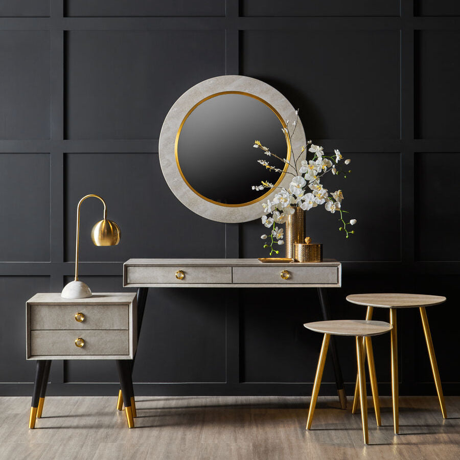 Dressing table with round mirror, lamp, and stools against a dark paneled wall.