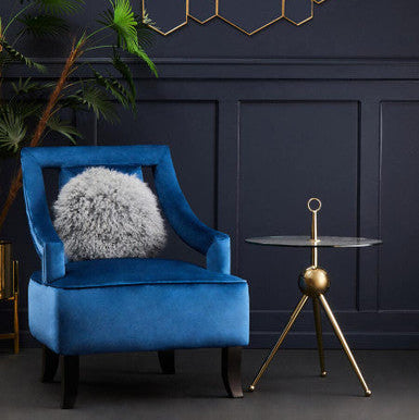 Blue armchair with a fluffy cushion in a room with dark walls, a geometric wall art piece, and a gold side table.