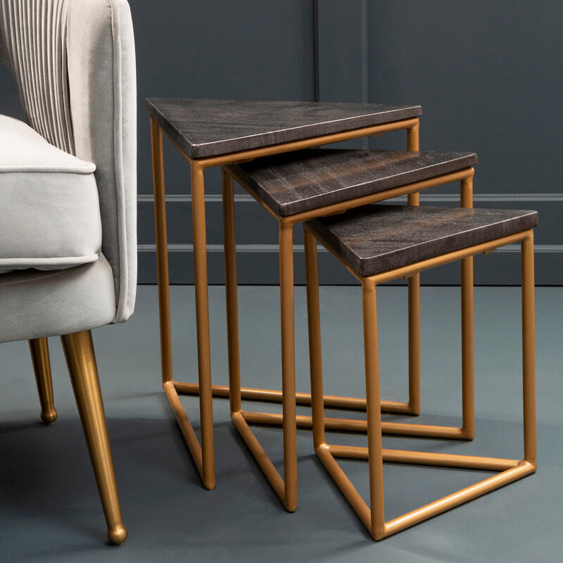 Set of three nesting tables with gold frames and dark wood tops next to a gray chair against a gray wall.