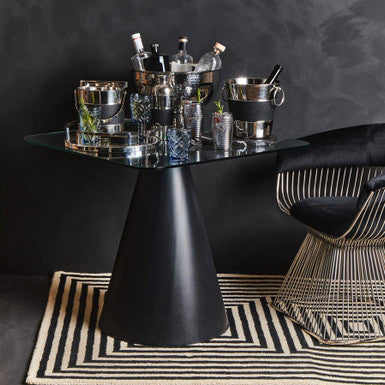 Modern black dining table with glass top in a room with dark walls and a patterned rug.