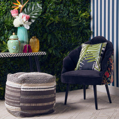 Navy chair with a floral pillow next to a patterned ottoman in a room with greenery and striped wall.