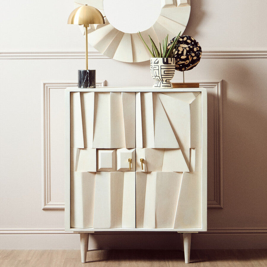 Decorative cabinet with abstract design, round mirror, and plants against a light wall.