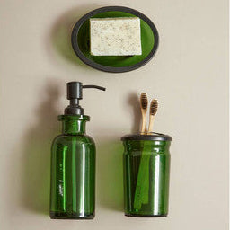 Green glass soap dispenser and jar with toothbrush holder on a beige wall.