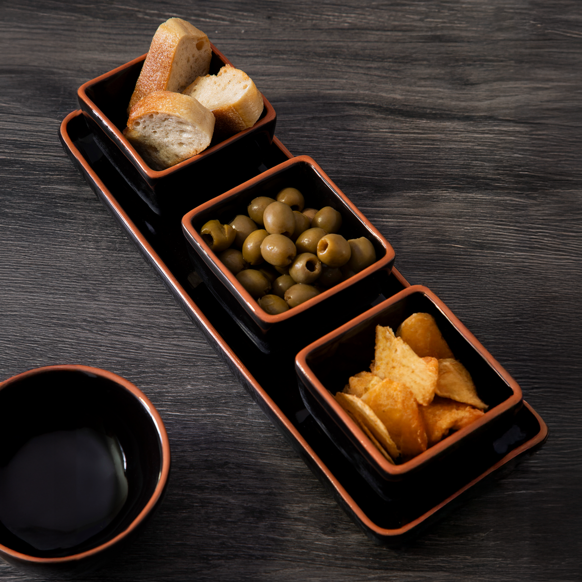 Three black ceramic dishes with bread, olives, and chips on a dark wooden surface.