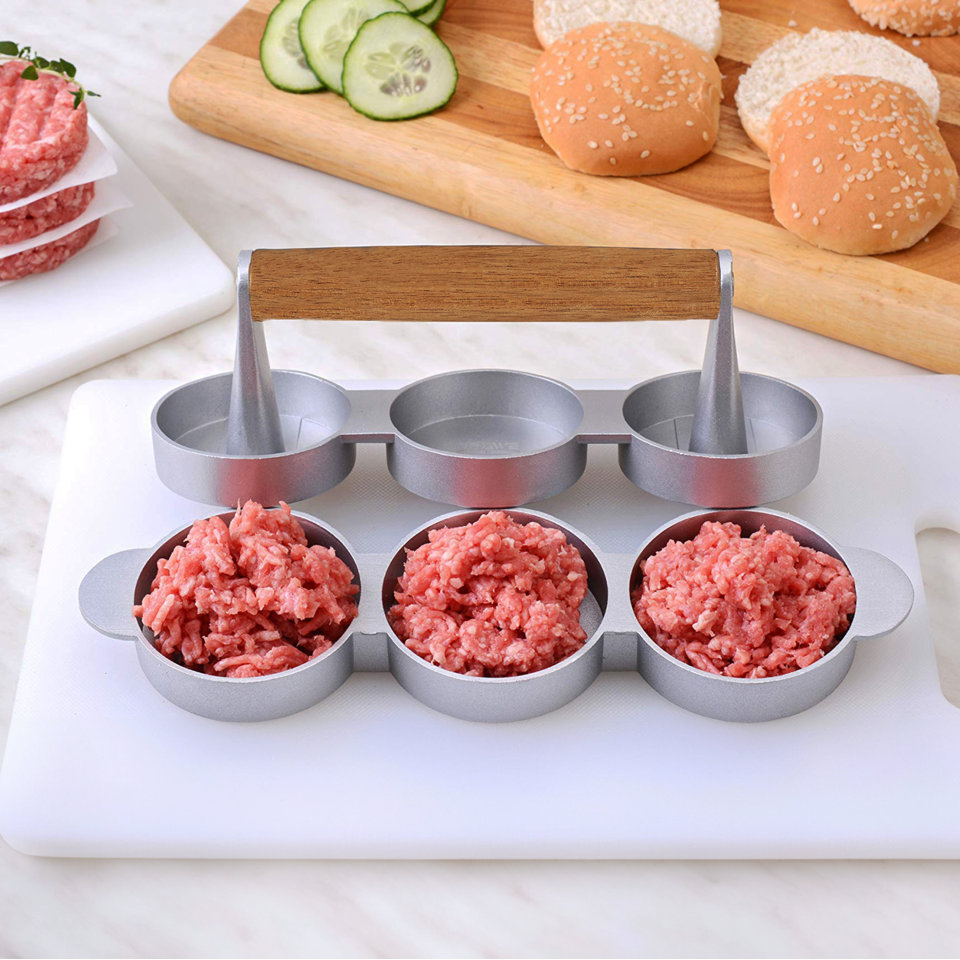 Hamburger meat forming tool with ground beef on a cutting board with buns and vegetables.