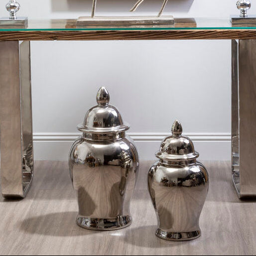 Decorative console table with silver vases, a glass top, and framed artwork on a white wall.