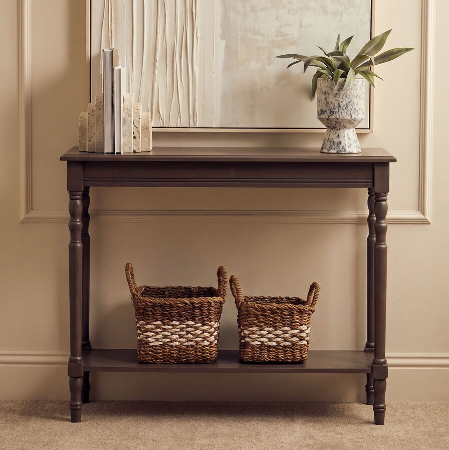 A dark brown wooden console table with a lower shelf, styled with two woven baskets, a small potted plant, and books, set against a beige paneled wall with a large mirror.