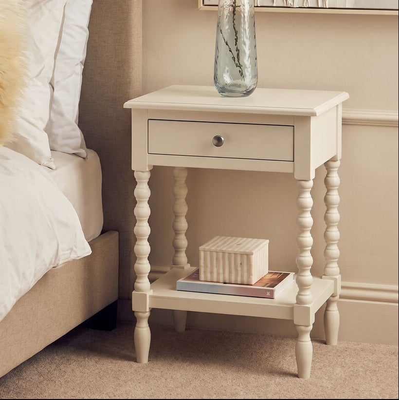 A cream-colored wooden bedside table with a single drawer and turned legs, styled next to a bed in a warm beige bedroom. A clear glass vase with greenery and a striped box sit on the table.