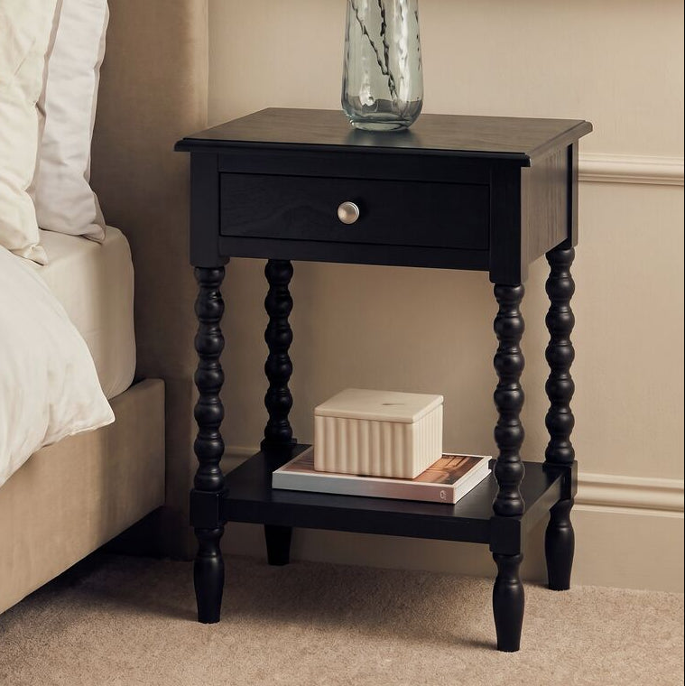 A black wooden bedside table with a single drawer and turned legs, styled next to a bed in a warm beige bedroom. A clear glass vase with eucalyptus stems sits on top, and a small box rests on the lower shelf.