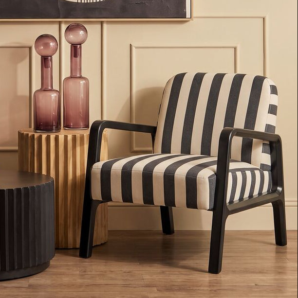 A styled interior scene with a black and white striped armchair, a black ribbed coffee table, pink glass bottles on a gold side table, and a large framed abstract wall art piece on a beige paneled wall.