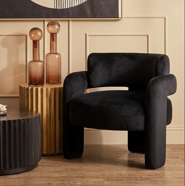 A styled interior scene with a black velvet armchair, a dark ribbed coffee table, decorative amber bottles on a gold side table, and a large abstract wall art piece above a beige paneled wall.