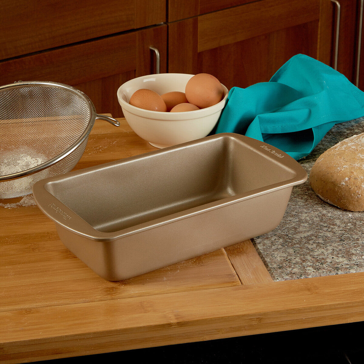Bread loaf pan on a kitchen counter with eggs, flour, and a teal towel.