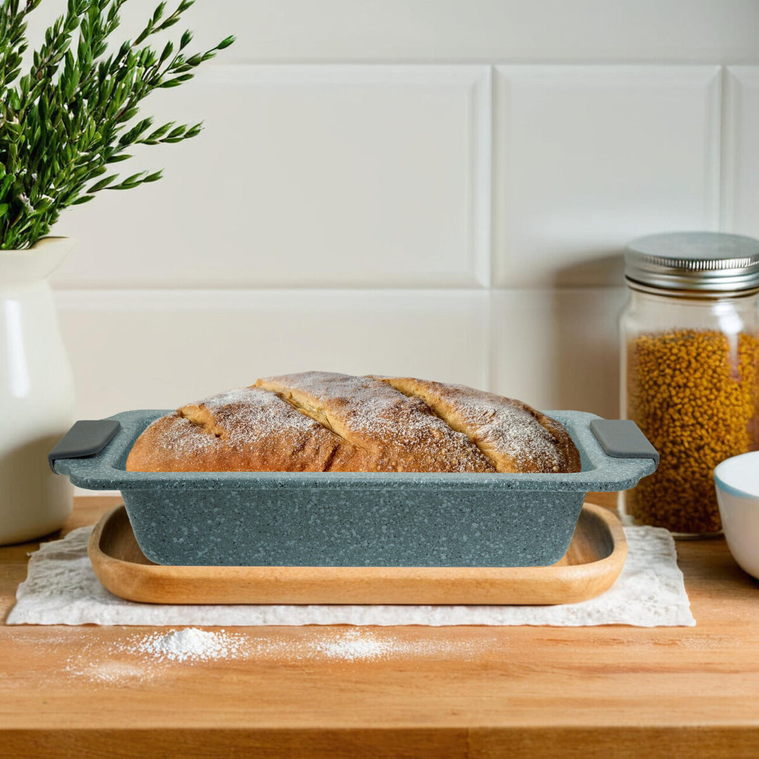 Loaf of bread in a textured loaf pan on a wooden surface with a plant and jars in the background.