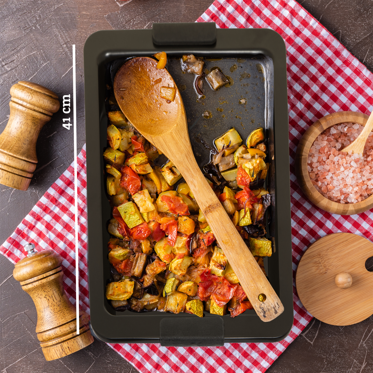 Roasted vegetables in a baking tray with a wooden spoon on a checkered tablecloth.
