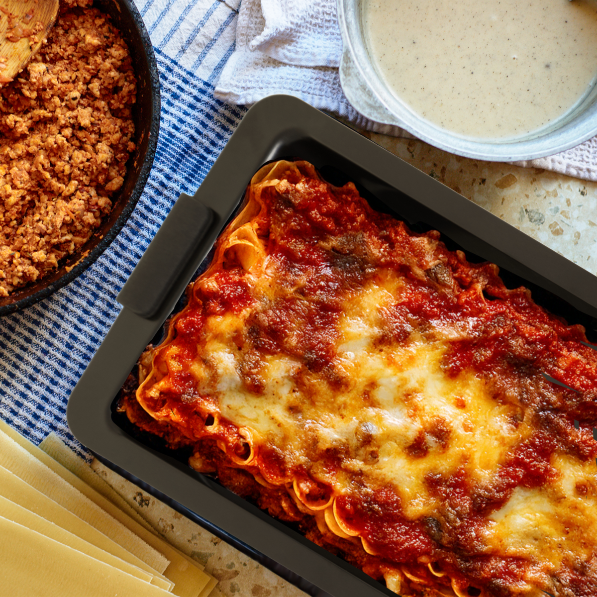 Baked lasagna in a black dish with ingredients like pasta and sauce on a blue checkered tablecloth.